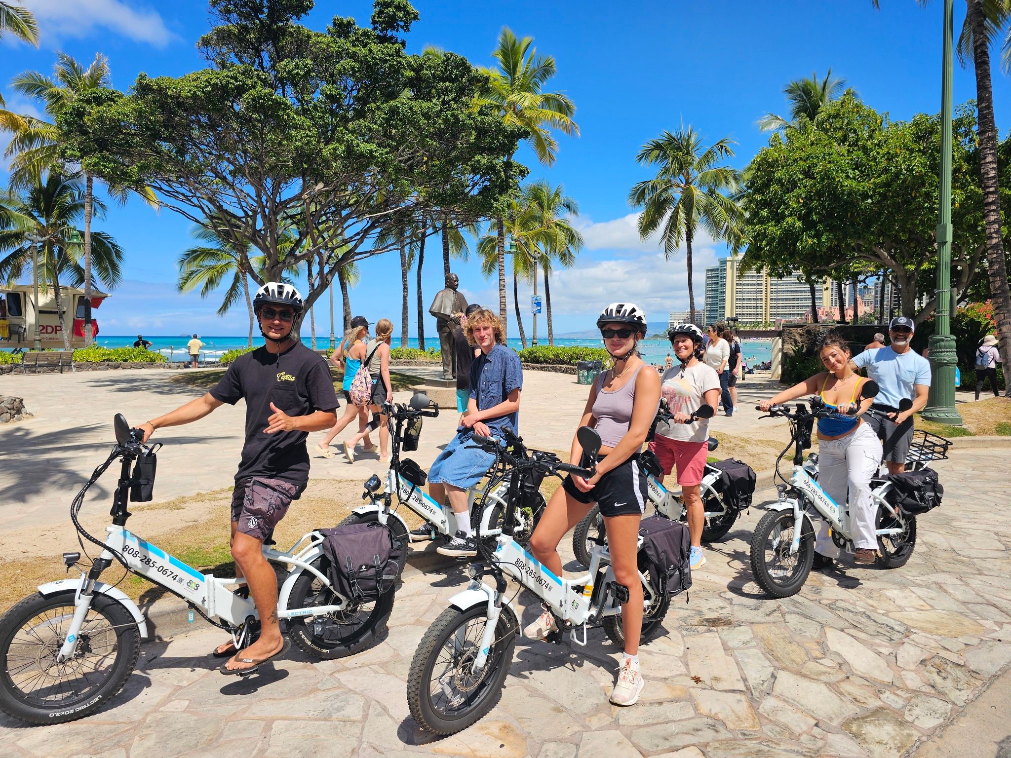 Group of people on electric bikes at a sunny beachfront in Honolulu, palm trees and turquoise ocean with hotel skyline in the background.