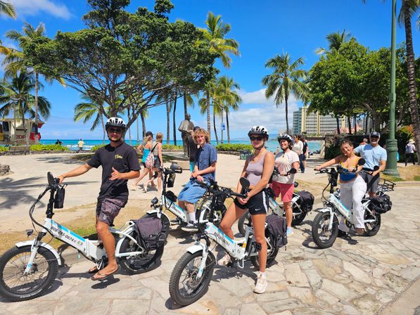Group of people on electric bikes at a sunny beachfront in Honolulu, palm trees and turquoise ocean with hotel skyline in the background.