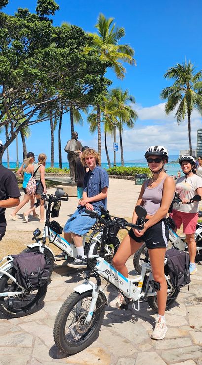 Group of people on electric bikes at a sunny beachfront in Honolulu, palm trees and turquoise ocean with hotel skyline in the background.