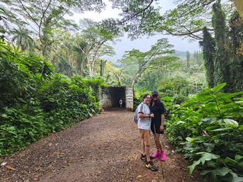 Two hikers posing on a muddy tropical rainforest trail beside dense green foliage and a graffiti-covered shipping container tunnel, with tall trees and misty hills in the background.