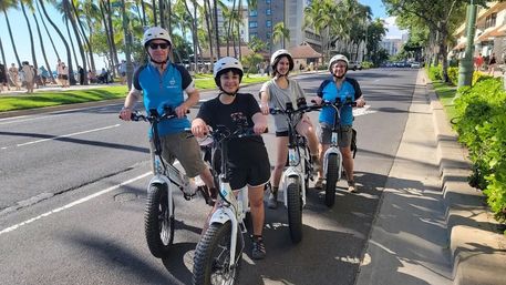 Four helmeted riders on white electric fat‑tire bikes cruising a palm‑lined coastal avenue beside a sunny beachfront promenade and city buildings.