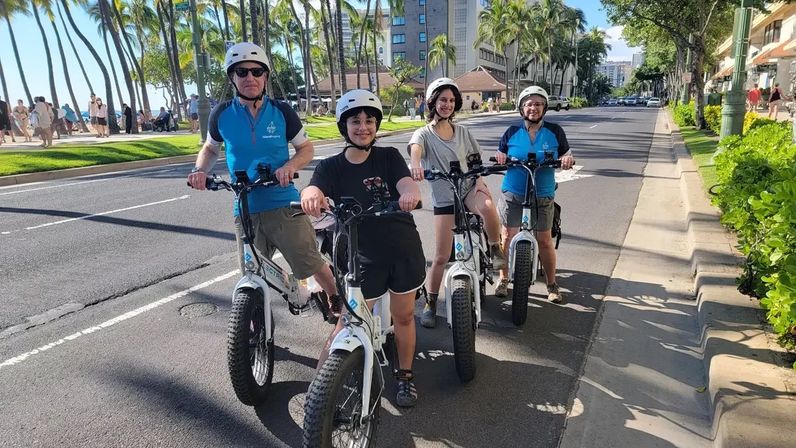 Four helmeted riders on white electric fat‑tire bikes cruising a palm‑lined coastal avenue beside a sunny beachfront promenade and city buildings.