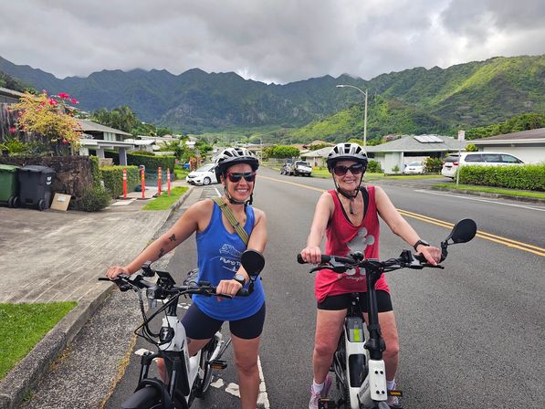 Two smiling cyclists in helmets on electric bikes pause on a residential street with lush green tropical mountains and a cloudy sky in the background.