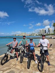 Four people wearing helmets on electric bikes along a sunny Waikiki beachfront, turquoise ocean and Honolulu skyline in the background.