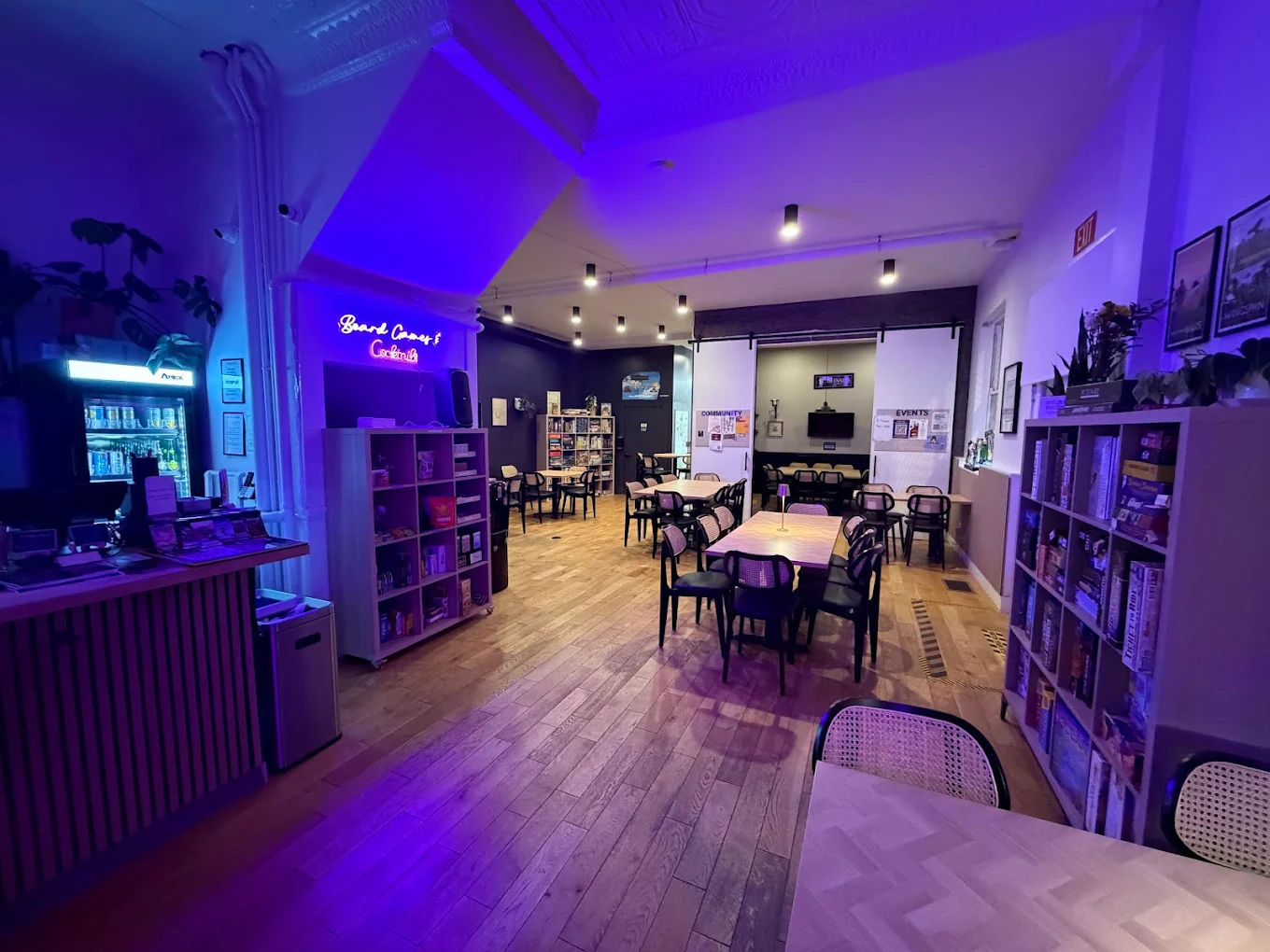 Cozy board game café interior with wooden floors, rows of tables and chairs, shelves stocked with board games, and purple-blue neon ambient lighting.