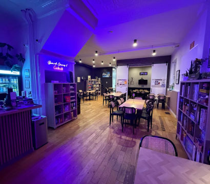 Cozy board game café interior with wooden floors, rows of tables and chairs, shelves stocked with board games, and purple-blue neon ambient lighting.