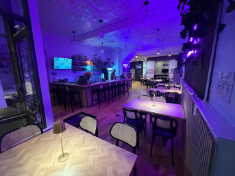 Neon-lit urban café-bar interior in purple-blue glow with herringbone wood tables and small lamps, rattan-back chairs, long bar with high stools, pendant lights and wall plants — cozy late-night dining vibe