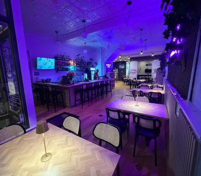 Neon-lit urban café-bar interior in purple-blue glow with herringbone wood tables and small lamps, rattan-back chairs, long bar with high stools, pendant lights and wall plants — cozy late-night dining vibe