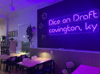 Cozy game-cafe interior bathed in purple neon light, with communal wooden tables, wicker-back chairs, bookshelf, hanging plants and warm table lamps.