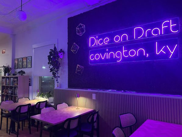 Cozy game-cafe interior bathed in purple neon light, with communal wooden tables, wicker-back chairs, bookshelf, hanging plants and warm table lamps.