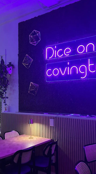 Cozy game-cafe interior bathed in purple neon light, with communal wooden tables, wicker-back chairs, bookshelf, hanging plants and warm table lamps.