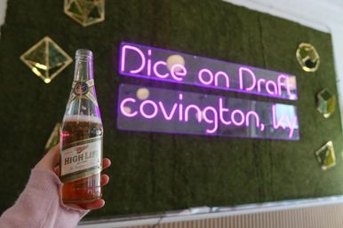 Hand holding a chilled Miller High Life beer bottle in front of a neon pink wall sign reading Covington, KY on a moss-covered backdrop