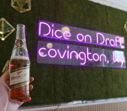 Hand holding a chilled Miller High Life beer bottle in front of a neon pink wall sign reading Covington, KY on a moss-covered backdrop
