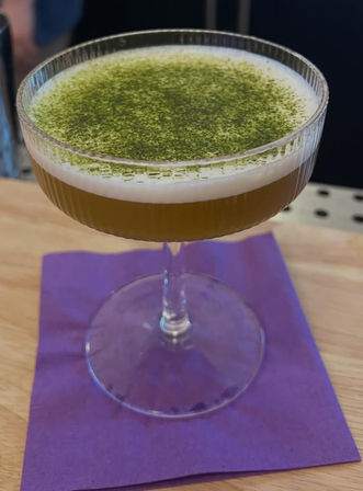 Matcha-topped craft cocktail in a coupe glass with creamy foam, served on a purple napkin at a wooden bar