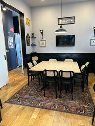 Cozy dining/meeting area with a square light wood table seating eight on a patterned rug, cane-back chairs, wood floors, black wainscoting, wall-mounted TV and hanging pendant light — casual urban bistro vibe.