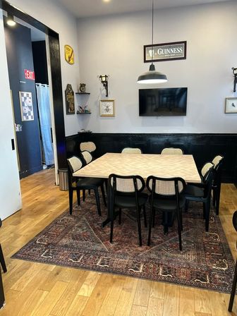 Cozy dining/meeting area with a square light wood table seating eight on a patterned rug, cane-back chairs, wood floors, black wainscoting, wall-mounted TV and hanging pendant light — casual urban bistro vibe.