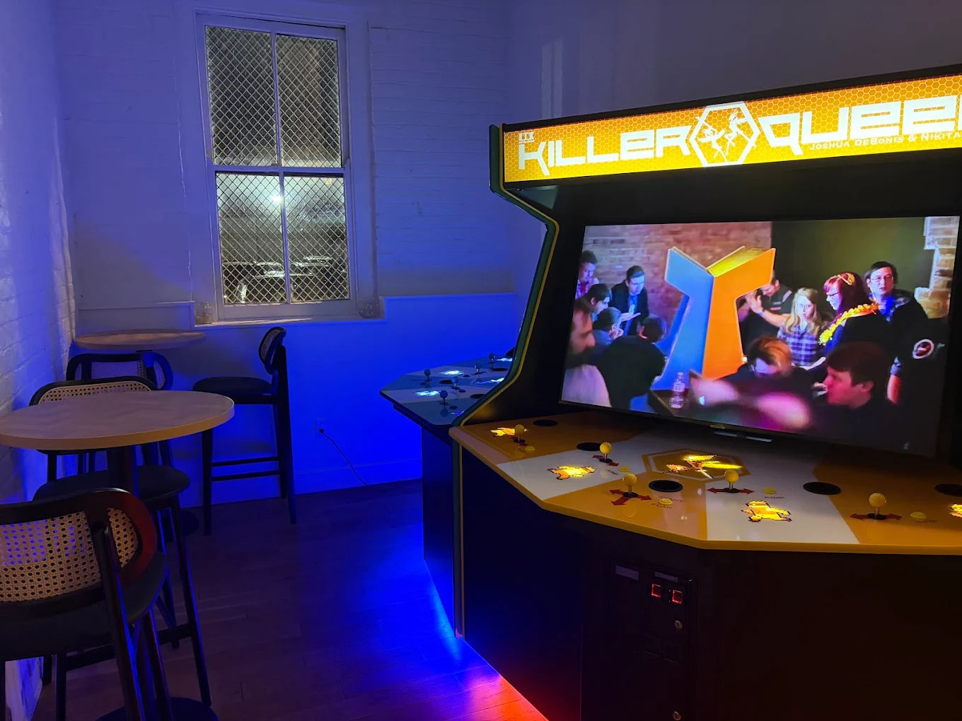 Retro multiplayer arcade cabinet with yellow marquee and crowd video on screen in a blue-lit gaming lounge with high-top tables and a wire-mesh window.