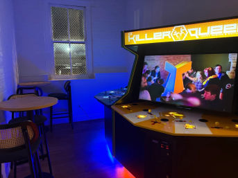 Retro multiplayer arcade cabinet with yellow marquee and crowd video on screen in a blue-lit gaming lounge with high-top tables and a wire-mesh window.