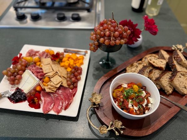Charcuterie spread on a home kitchen island: plate of sliced salami, brie with jam, cubed cheddar, crackers, grapes and cherry tomatoes; pedestal bowl of red grapes, bowl of roasted cherry tomatoes with herbs and cheese, and sliced toasted bread on a wooden board.