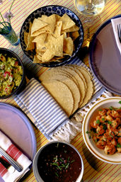 Overhead shot of a Mexican-inspired table spread: bowl of tortilla chips, stacked corn tortillas on a striped napkin, guacamole, black beans and seasoned shrimp on colorful plates.