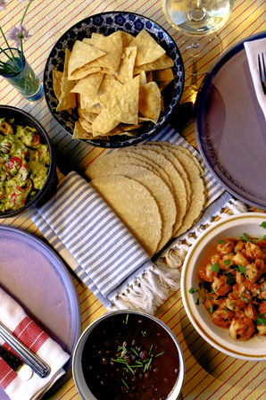 Overhead shot of a Mexican-inspired table spread: bowl of tortilla chips, stacked corn tortillas on a striped napkin, guacamole, black beans and seasoned shrimp on colorful plates.
