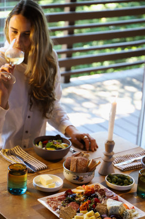 Person sipping white wine at a cozy candlelit patio on a wooden deck, with a charcuterie board, olives, bread and salad for relaxed outdoor dining