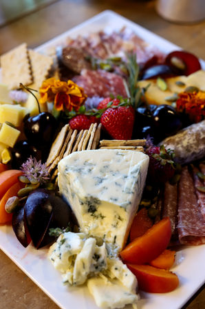 Close-up gourmet charcuterie board with blue cheese wedge, cubed cheddar, crackers, strawberries, dark grapes, plum slices, salami and edible flowers