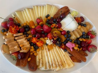 White oval platter with a colorful cheese and fruit grazing board: fanned slices of semi-hard cheese, a soft goat cheese log, crackers and crostini, strawberries, cherries, blueberries, dried apricots, crunchy nuts and edible flowers.