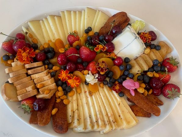 White oval platter with a colorful cheese and fruit grazing board: fanned slices of semi-hard cheese, a soft goat cheese log, crackers and crostini, strawberries, cherries, blueberries, dried apricots, crunchy nuts and edible flowers.