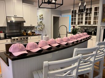 Playful row of pastel pink baseball caps displayed on a black granite kitchen island in a bright modern white home kitchen with white bar stools, stainless-steel range, and fresh flowers.