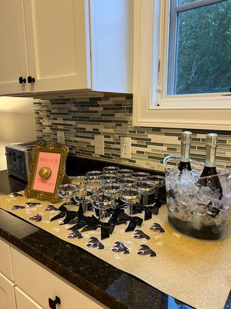 Home kitchen countertop party setup with rows of champagne coupes tied with black bows on a gold runner, ice bucket holding chilled champagne bottles, and a framed “Ring for Champagne” bell by a mosaic tile backsplash.