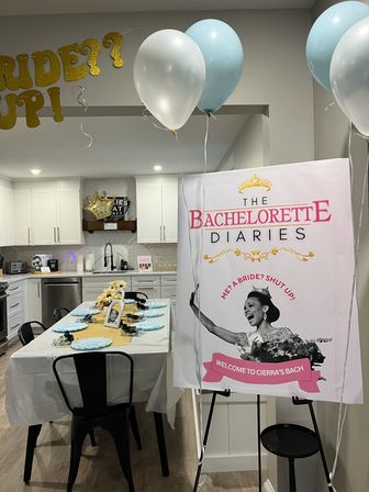 Bachelorette party setup in a modern home kitchen with gold, white and blue balloons, a themed “Bachelorette Diaries” welcome poster, and a decorated dining table