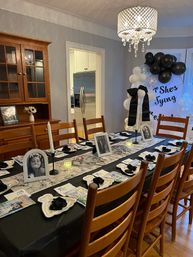 Black-and-white themed home dining room party setup with a long table dressed with framed photos, candles, napkins, gift bags, a balloon arch and crystal chandelier, kitchen visible in the background.