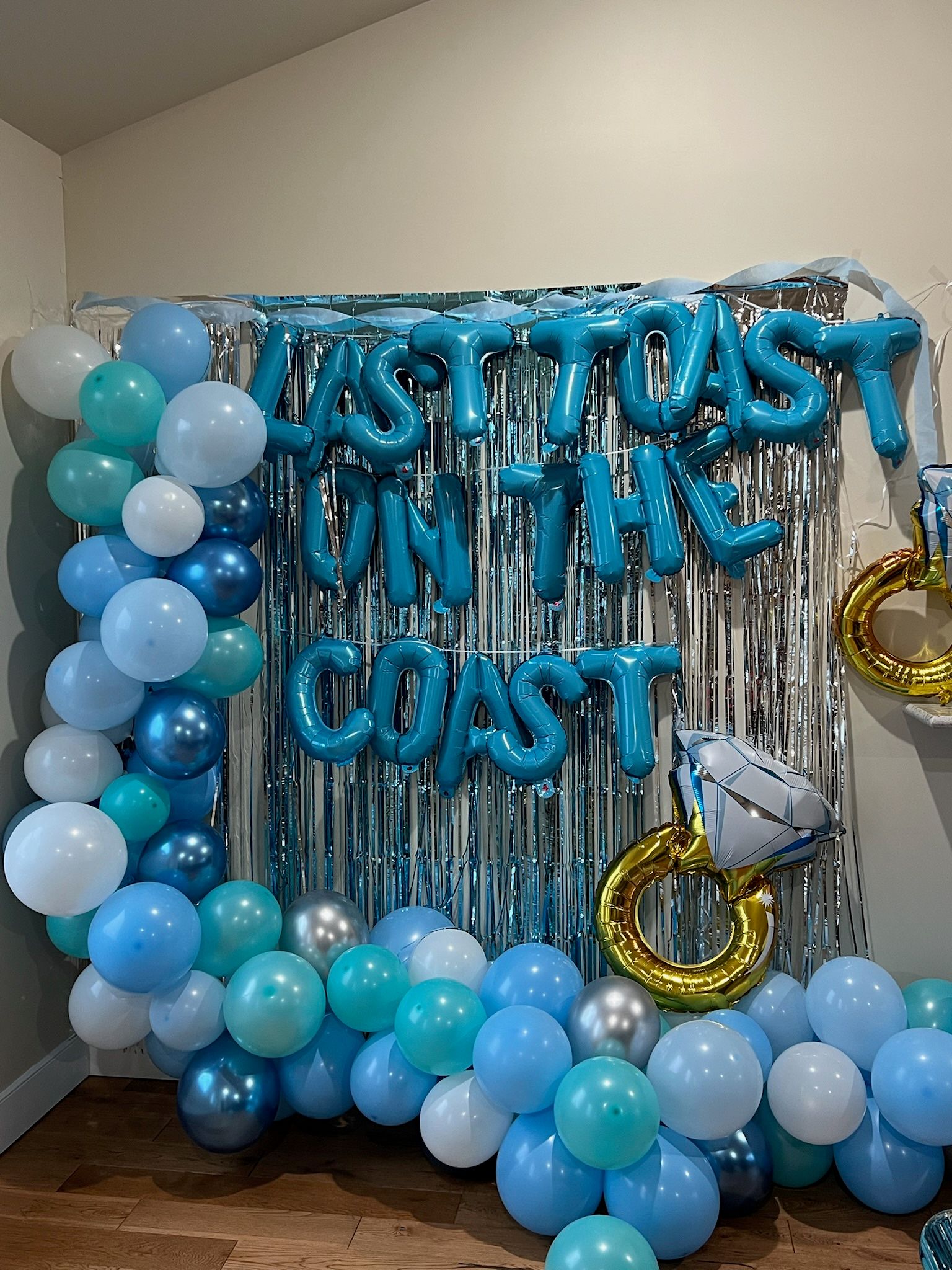 Silver fringe backdrop decorated with teal, aqua, sky-blue, white and metallic balloons, blue letter foil balloons spelling a celebratory message, and a gold ring-shaped foil balloon with a diamond — festive engagement or party backdrop.
