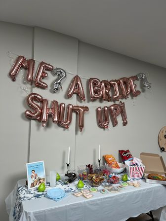 Rose-gold letter balloons reading "ME? A BRIDE? SHUT UP!" over an indoor snack table set for a bridal shower/bachelorette with candy bowls, cookies, chips, popcorn, candles and decorative pears.