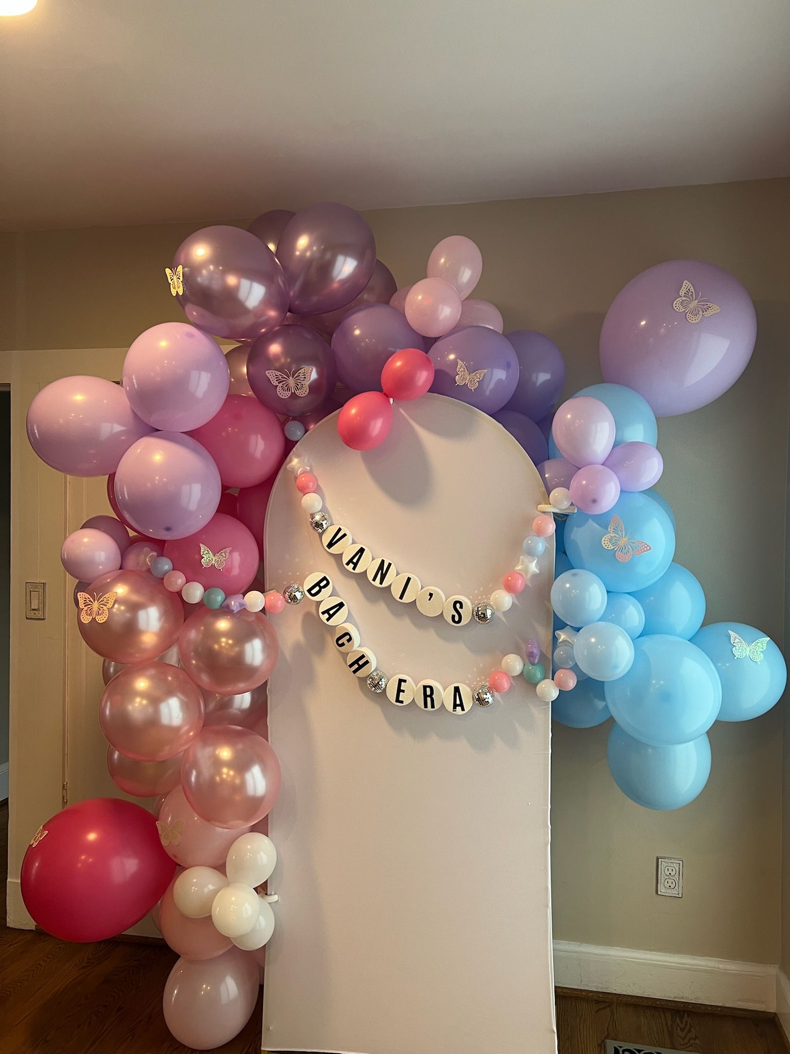 Playful pastel balloon arch in pink, lavender and blue with metallic accents and butterfly stickers framing a white display board, finished with round bead-style letter garlands for a celebratory backdrop.