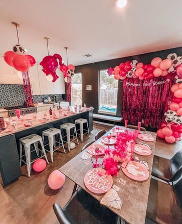 Barbie-themed bachelorette party setup in a modern home kitchen and dining area with pink and cow-print balloon garlands, metallic pink fringe backdrop reading “Barbie’s Last Rodeo,” barstool counter, themed place settings with pink feather boas and scattered balloons.
