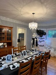 Home dining room decorated for a black-and-white celebration: long table with black tablecloth and white runner, framed black-and-white portraits, candles and place settings, crystal chandelier casting patterned light, black-and-white balloon arch by a bay window, wooden chairs and hardwood floor.