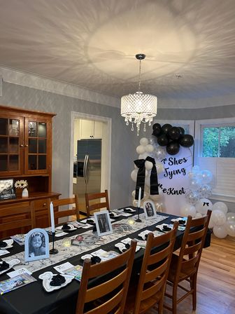 Home dining room decorated for a black-and-white celebration: long table with black tablecloth and white runner, framed black-and-white portraits, candles and place settings, crystal chandelier casting patterned light, black-and-white balloon arch by a bay window, wooden chairs and hardwood floor.