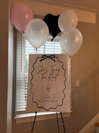 Playful indoor bachelorette welcome sign on an easel in a home entryway, surrounded by pink, white and black balloons; sign features a bow frame, script "She's Tying The Knot" and a small champagne glass icon.