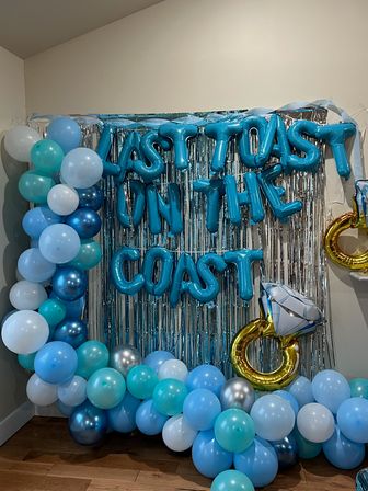 Blue and teal balloon garland framing a silver fringe backdrop with blue foil letter balloons forming a celebration phrase and a gold diamond-ring foil balloon — festive photo‑booth setup.