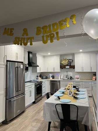 Open modern white kitchen decorated for a bridal shower with a gold glitter banner reading 'BRIDE?', a white balloon, and a long table set with blue plates, sunflower centerpiece and framed photos.