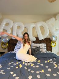 Bride-to-be in a white dress sitting on a bed under large white 'BRIDE' balloons with faux rose petals and LED candles for an indoor bridal shower celebration.