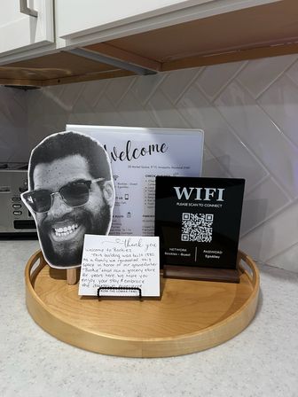 Kitchen countertop welcome tray with a round wooden tray holding a welcome sign, large black-and-white smiling face cutout, a handwritten guest note, and a black WiFi sign with QR code for network access.