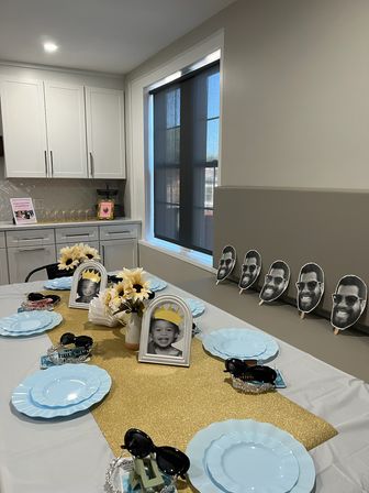 Modern home kitchen with a kids' party table: gold glitter runner, light blue plates, framed black-and-white child photos with paper crowns, sunflower centerpieces, sunglasses favors and repeated smiling face cutouts along the wall.