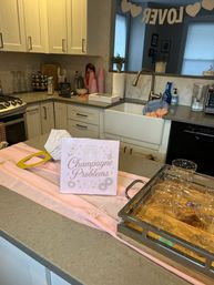 Pink-themed brunch setup on a modern home kitchen island with a “Champagne Problems” sign, champagne coupe glasses on a wooden tray, pink cups and paper decorations, farmhouse sink and a LOVE banner in the background