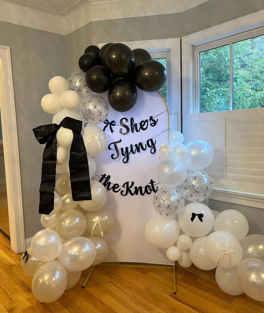 Indoor bridal shower backdrop with round white sign reading "She's Tying the Knot" framed by black, white and pearl balloon garlands, black ribbon bows, bay windows and hardwood floor