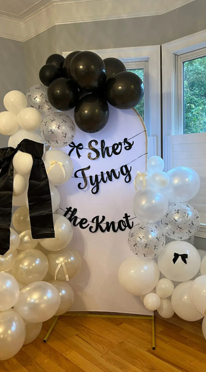 Indoor bridal shower backdrop with round white sign reading "She's Tying the Knot" framed by black, white and pearl balloon garlands, black ribbon bows, bay windows and hardwood floor