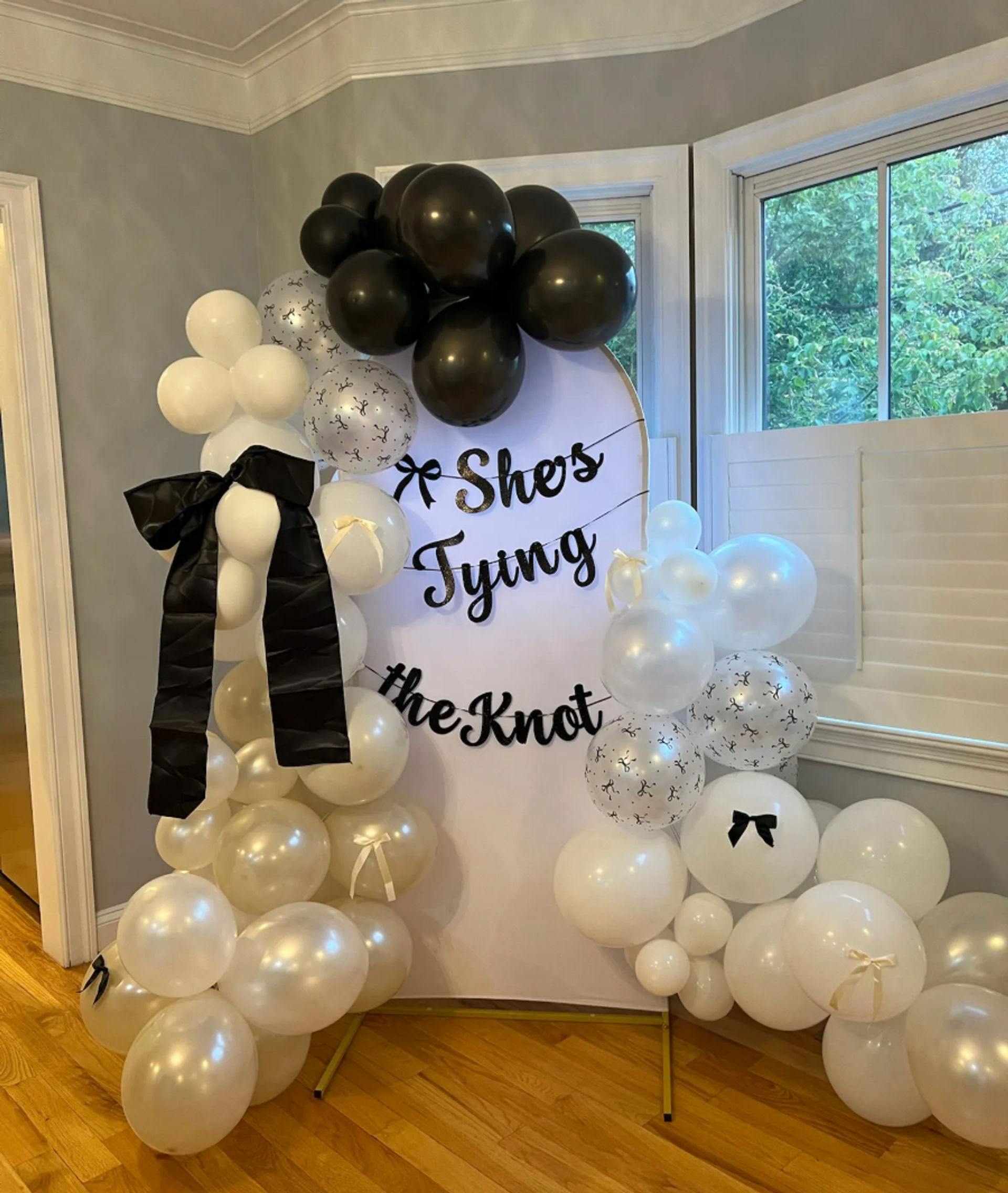 Indoor bridal shower backdrop with round white sign reading "She's Tying the Knot" framed by black, white and pearl balloon garlands, black ribbon bows, bay windows and hardwood floor