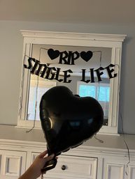 Hand holding a black heart-shaped balloon in front of a mirror decorated with a black "RIP SINGLE LIFE" banner, white dresser and window reflection — playful bachelorette party decor.